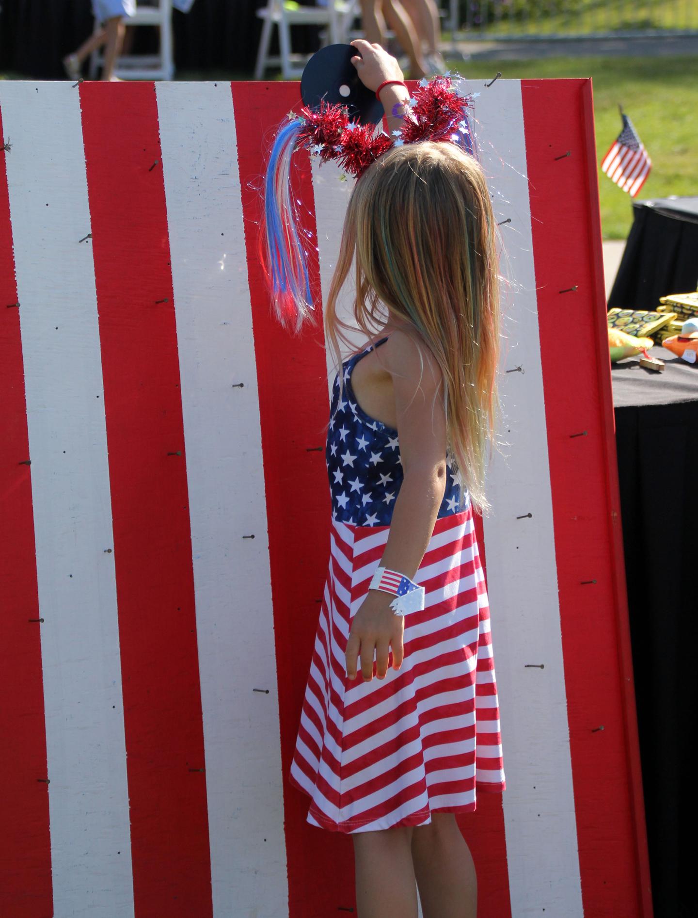 Child in a USA-themed dress reaching up, in front of a red and white striped backdrop.