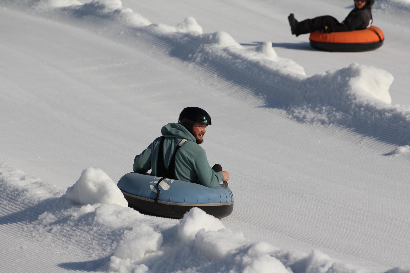 Two people snow tubing downhill on a snowy slope, one in blue, one in orange.