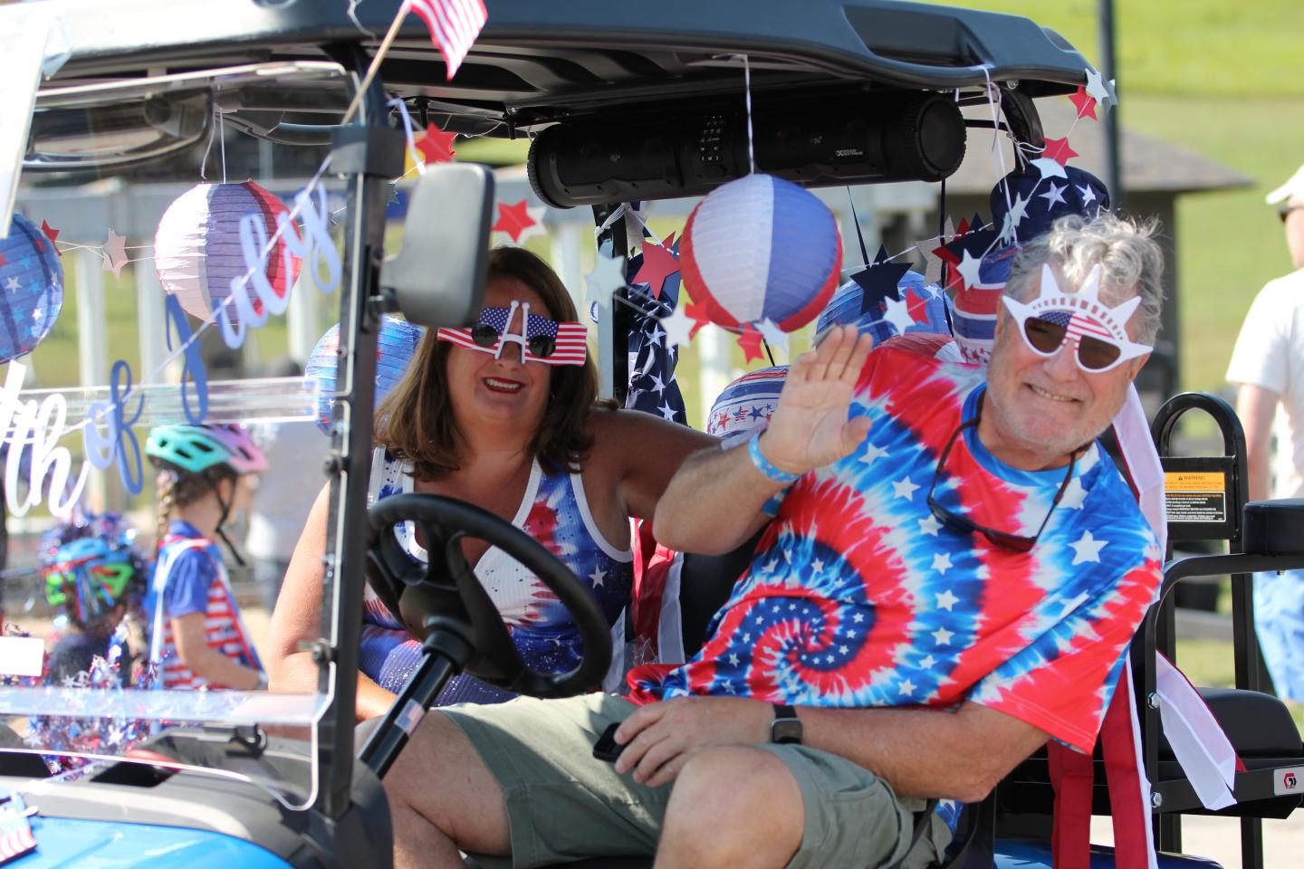 A couple in red, white, and blue outfits waves from a decorated golf cart.