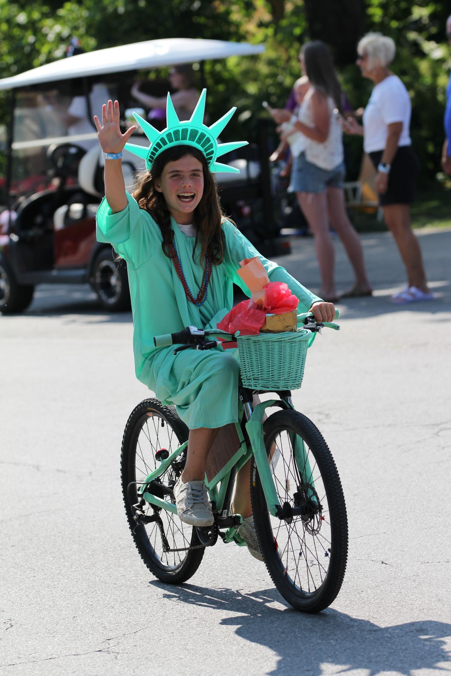 Child in Statue of Liberty costume riding a bike with a waving hand.