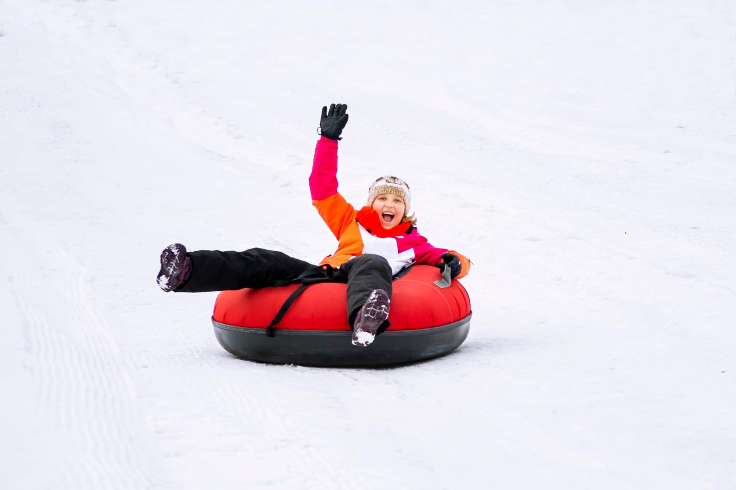 Child snow tubing in a bright red tube, smiling and waving.