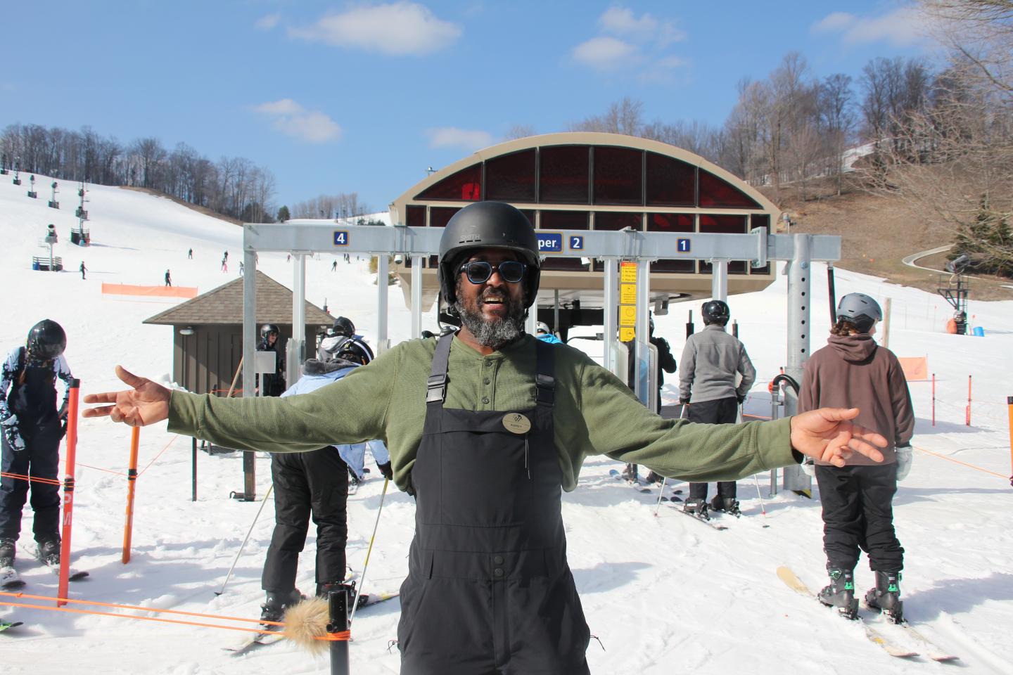 Man in ski gear smiling at a snowy ski resort with a lift in the background.
