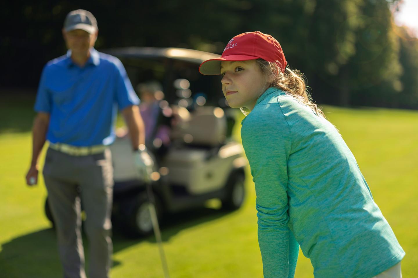 Young girl in a red cap playing golf, golf cart and man in the background.