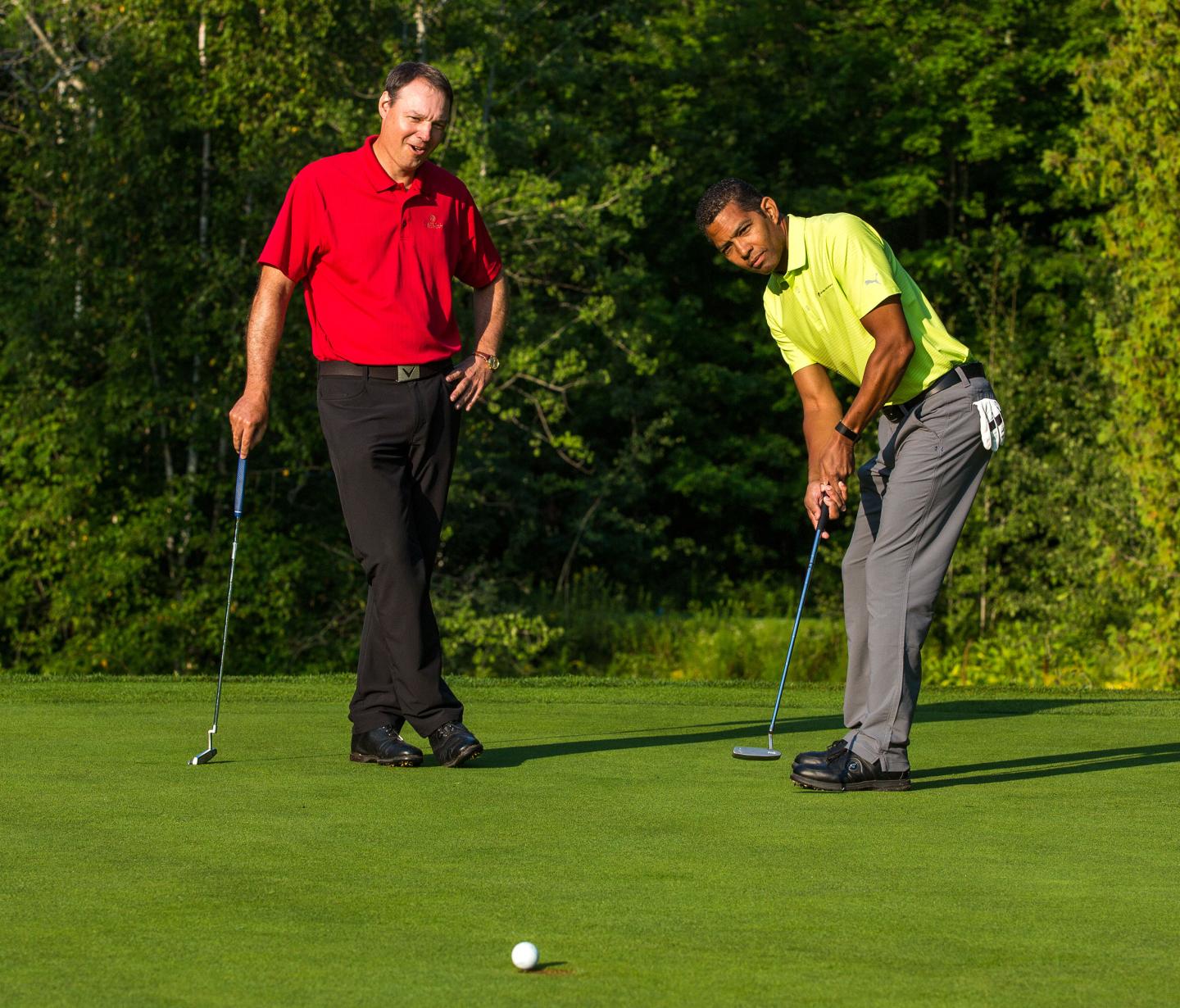 Two men golfing on a green, one in mid-putt, surrounded by trees.