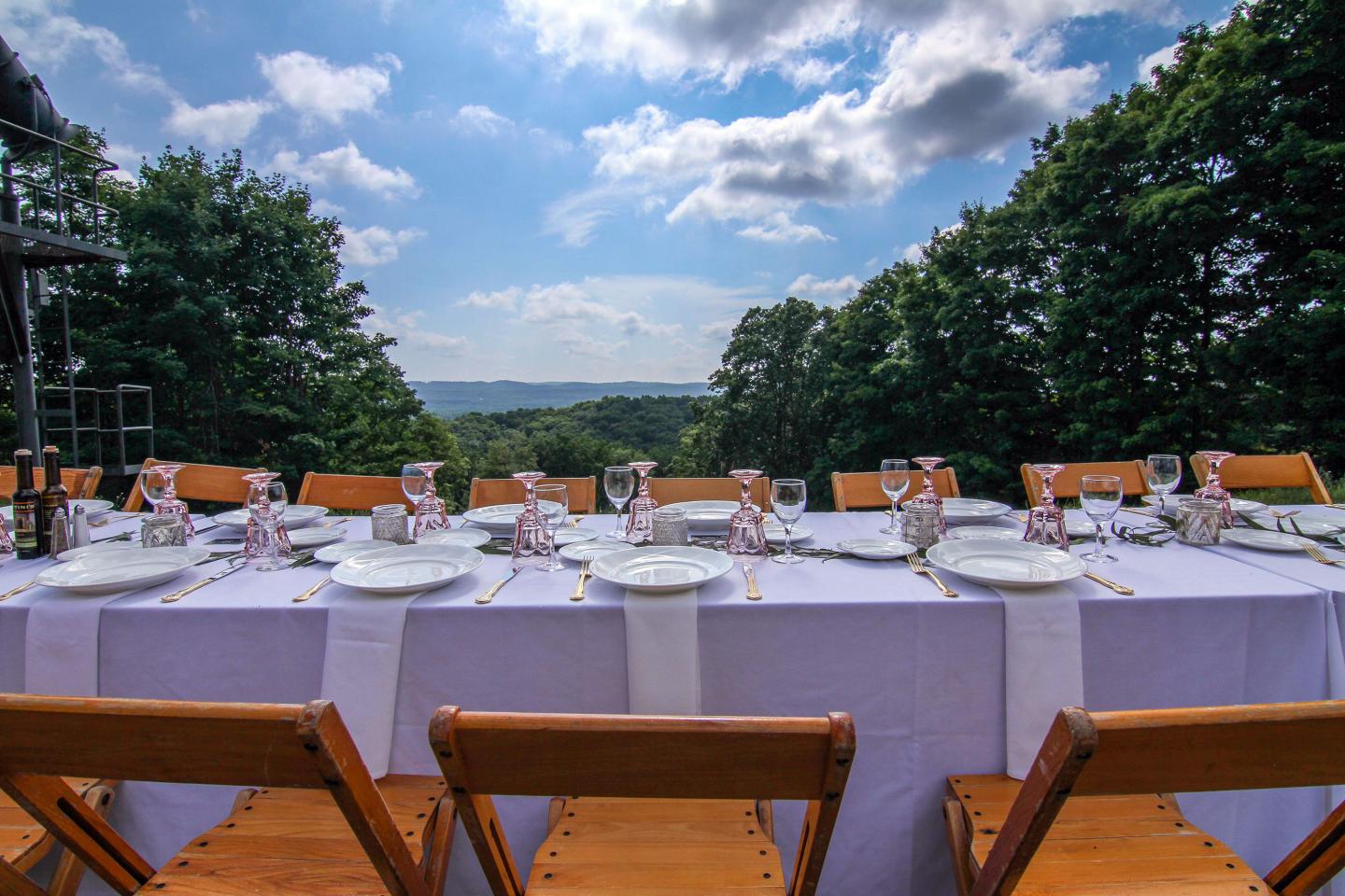 Outdoor dining table set with white linens, against a scenic backdrop of trees and a blue sky.