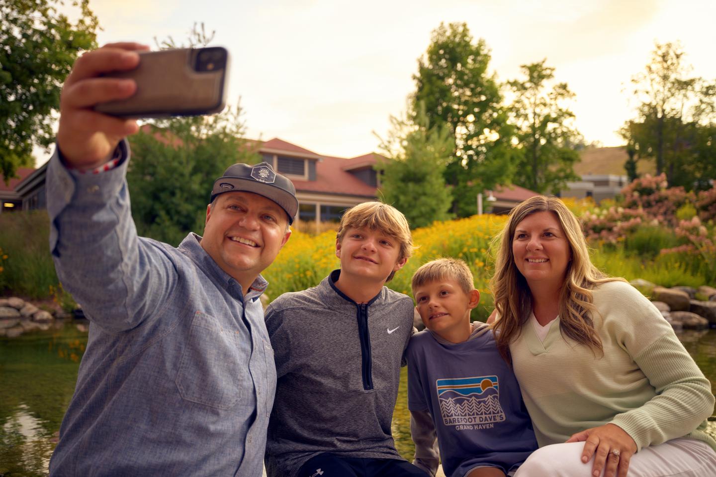 Family taking a selfie by a pond, smiling in casual clothes.