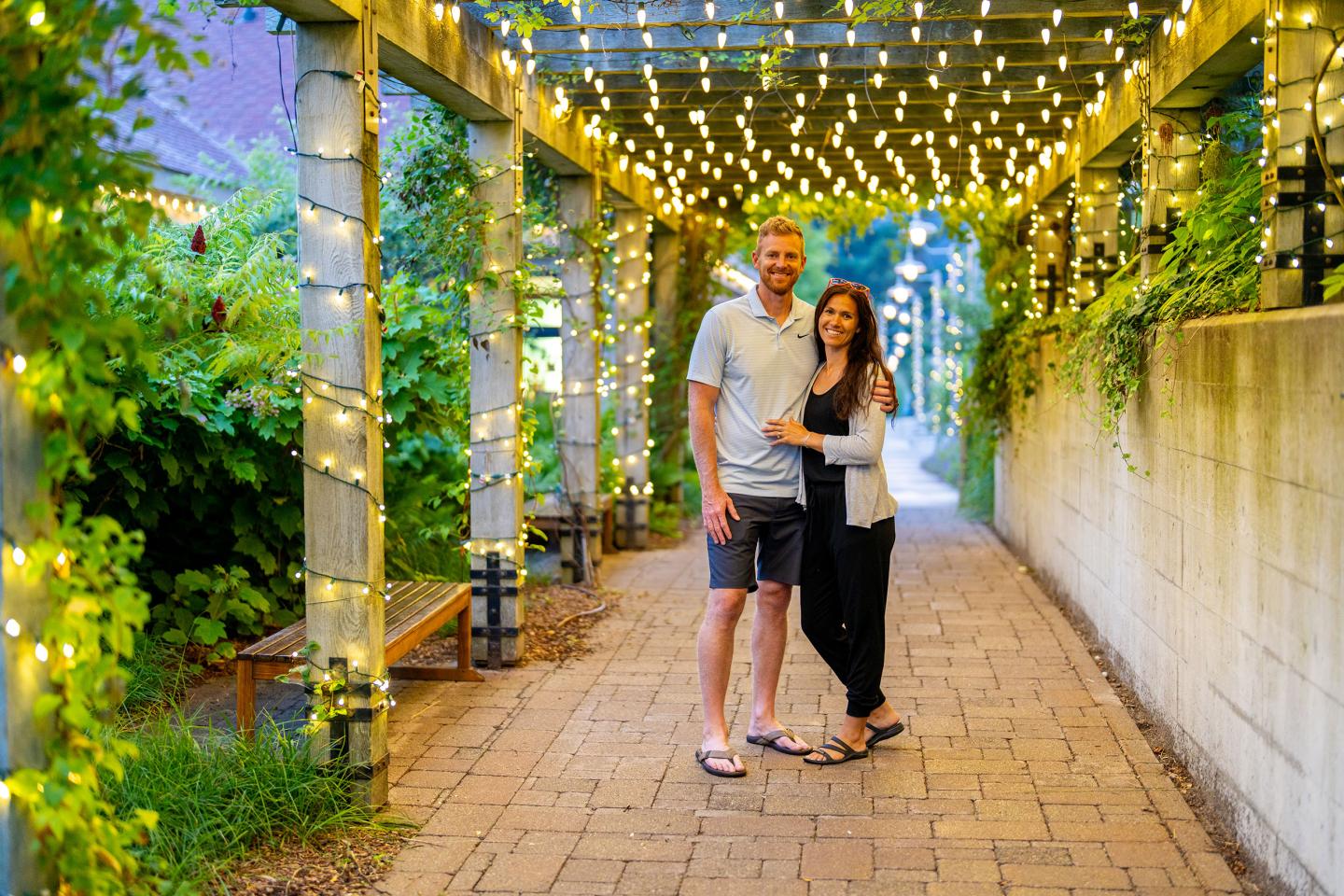 Couple smiling under string lights on a garden path.