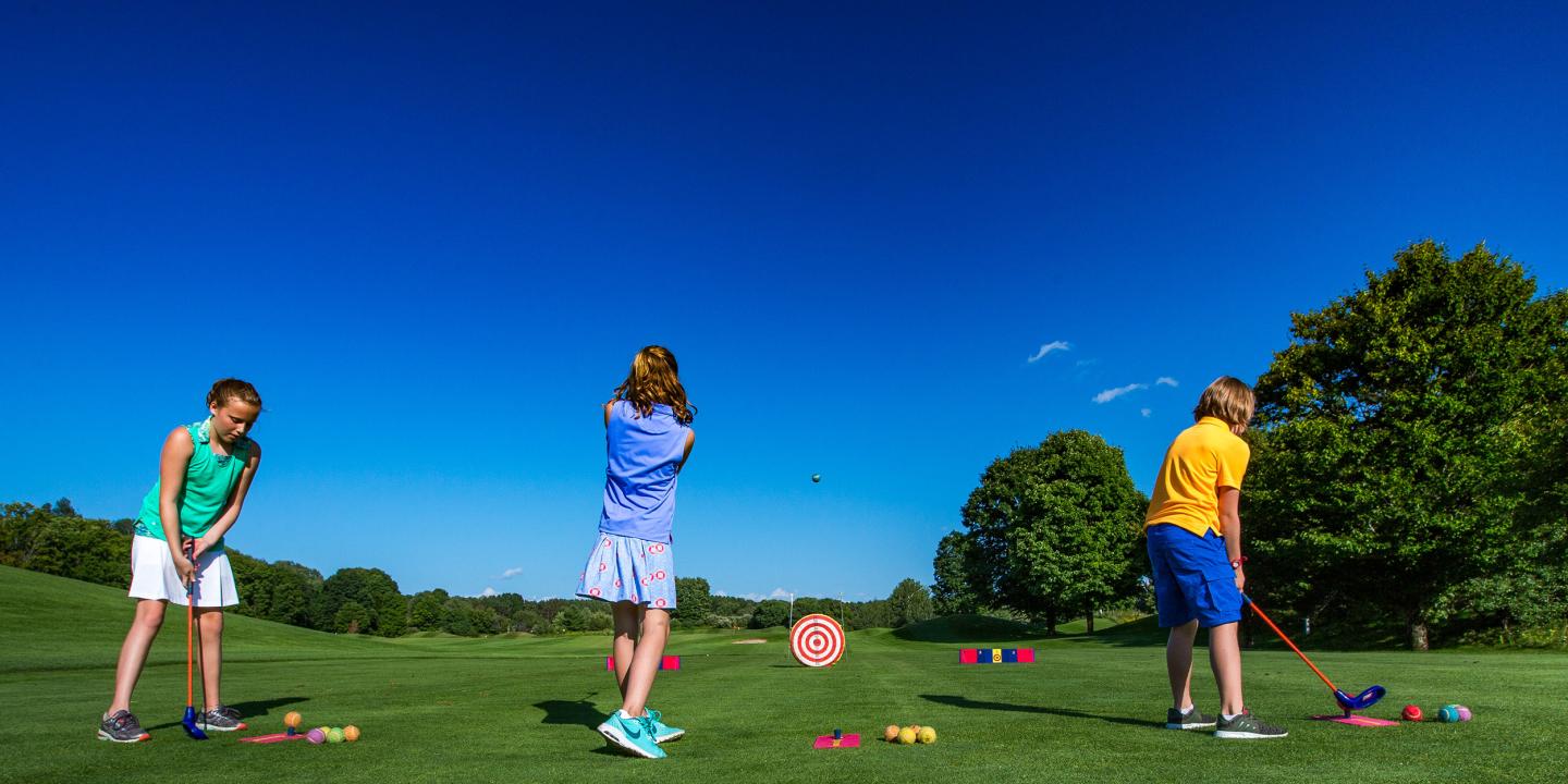 Kids playing golf on a grassy course under a clear blue sky.