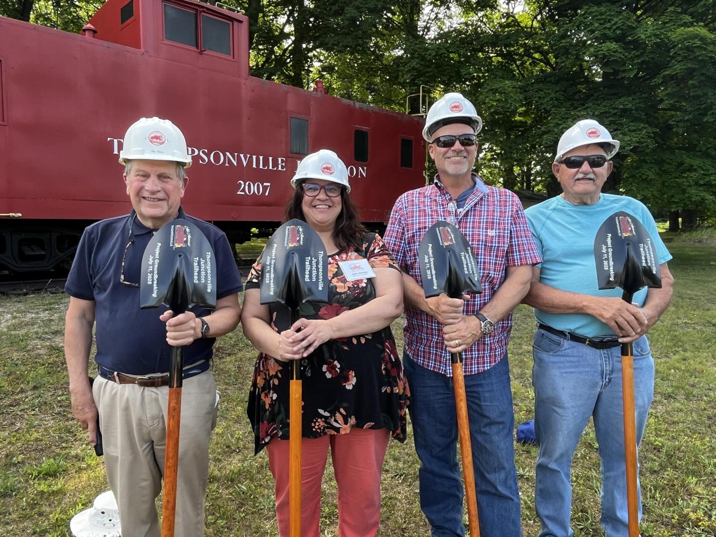Four people in hard hats holding shovels, standing in front of a red train car.