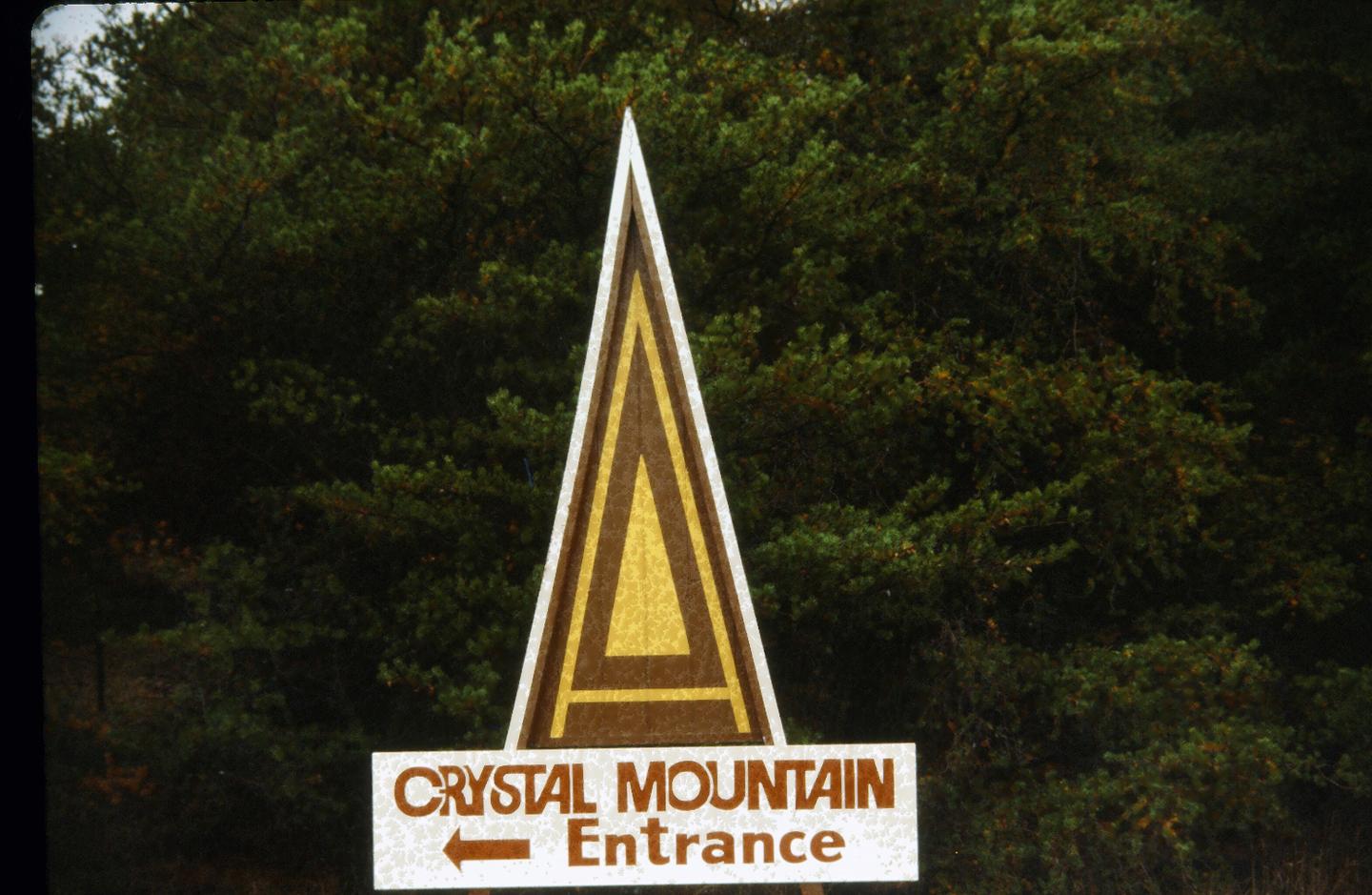 Triangular sign with "Crystal Mountain Entrance" in front of trees.
