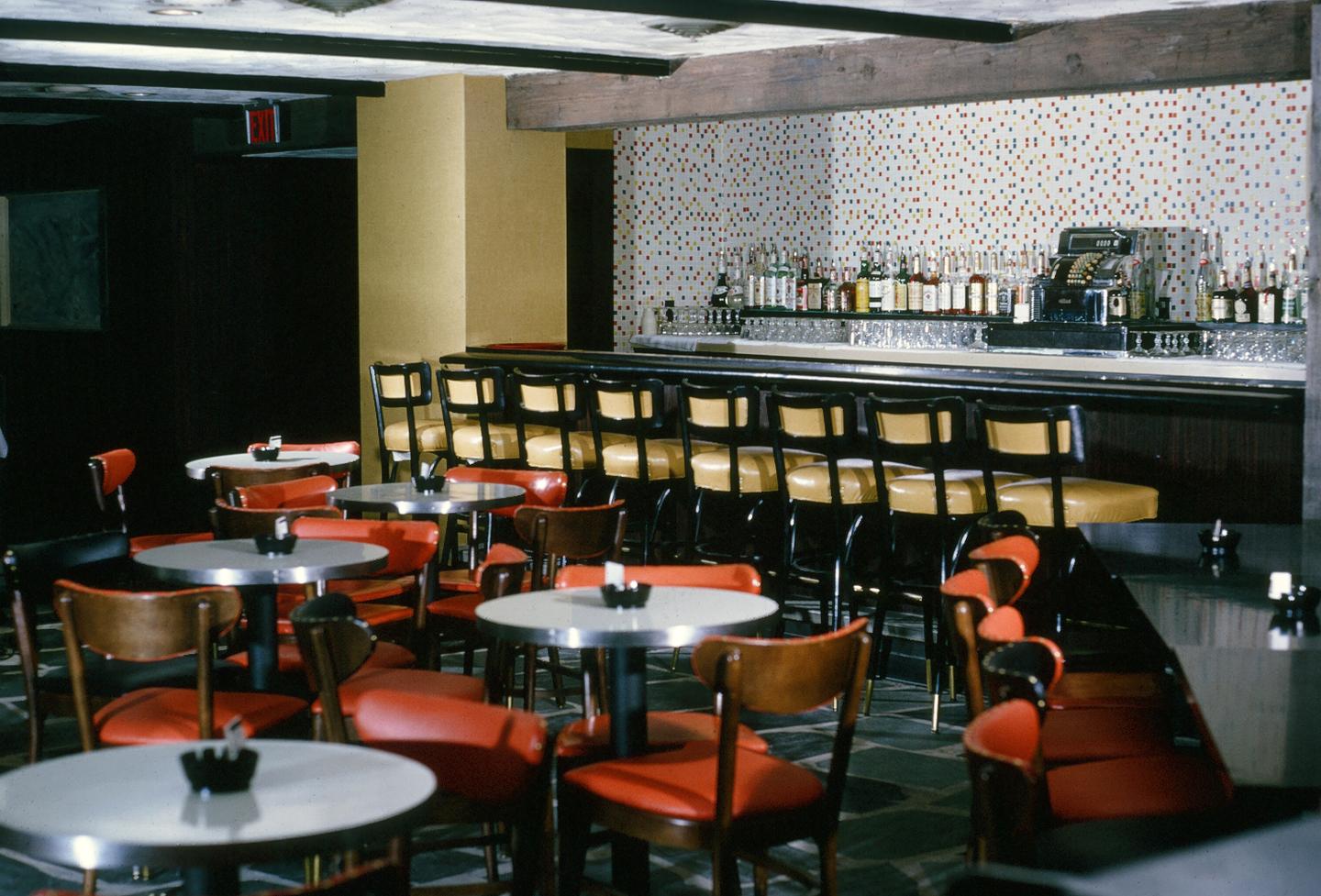 Empty bar with red and black seating, bottles on shelves, and a patterned wall.
