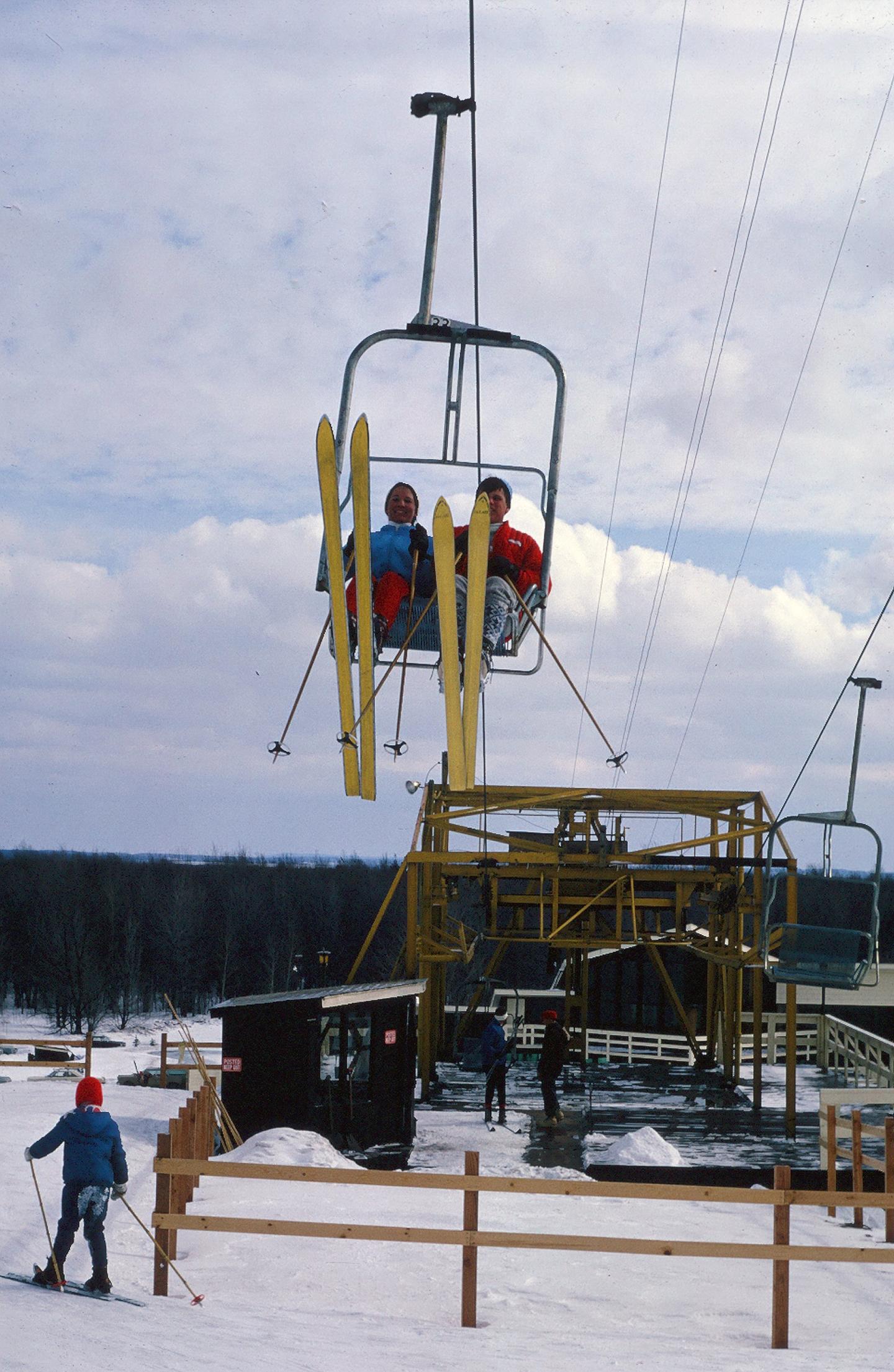 Skiers on a chairlift above a snowy slope, clear sky in the background.
