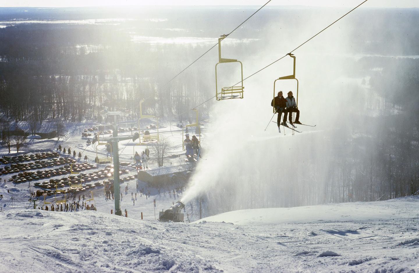 Ski lift ascending snowy slope, two people riding, snow cannon active nearby.