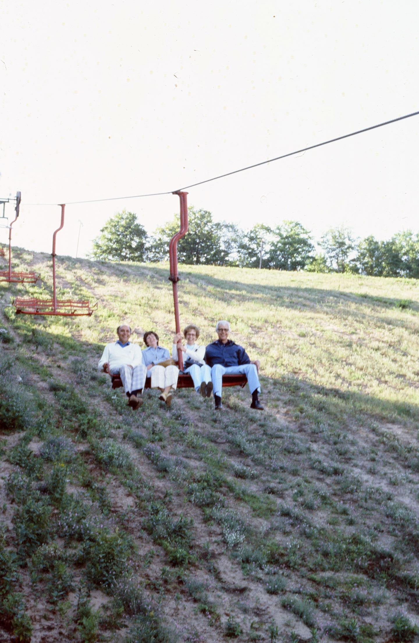 Four people sitting on a grassy hill, under a ski lift, with trees in the background.