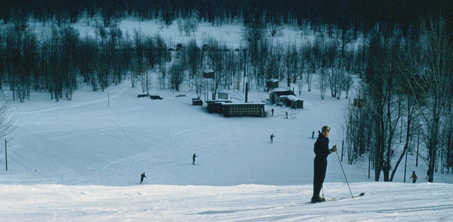 Snowy landscape with a skier on a hill and distant cabins.