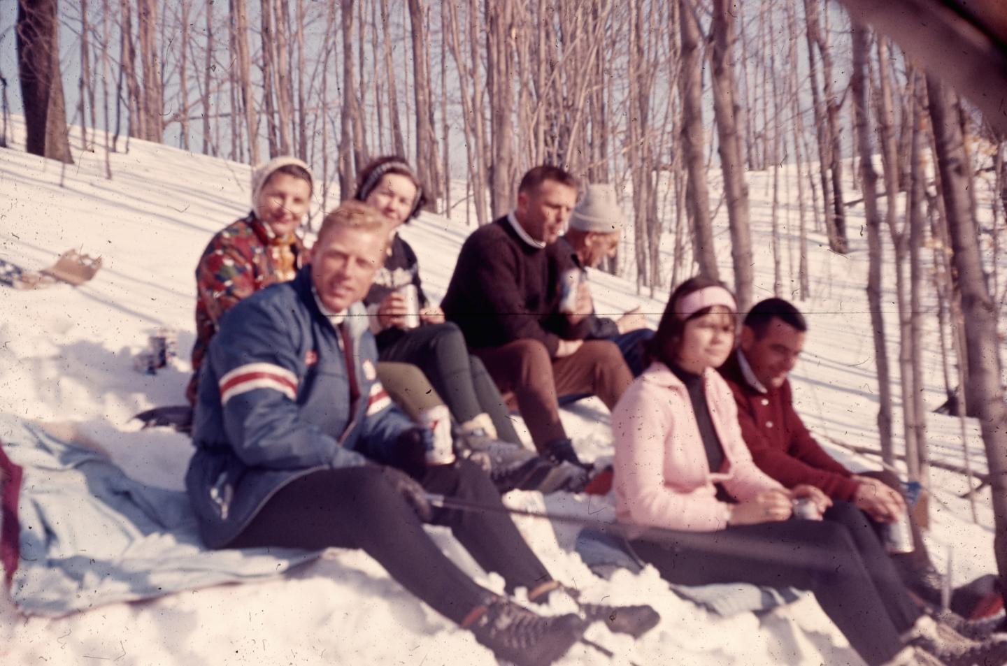 Group of people sitting on snowy hill, wearing warm clothes, surrounded by bare trees.