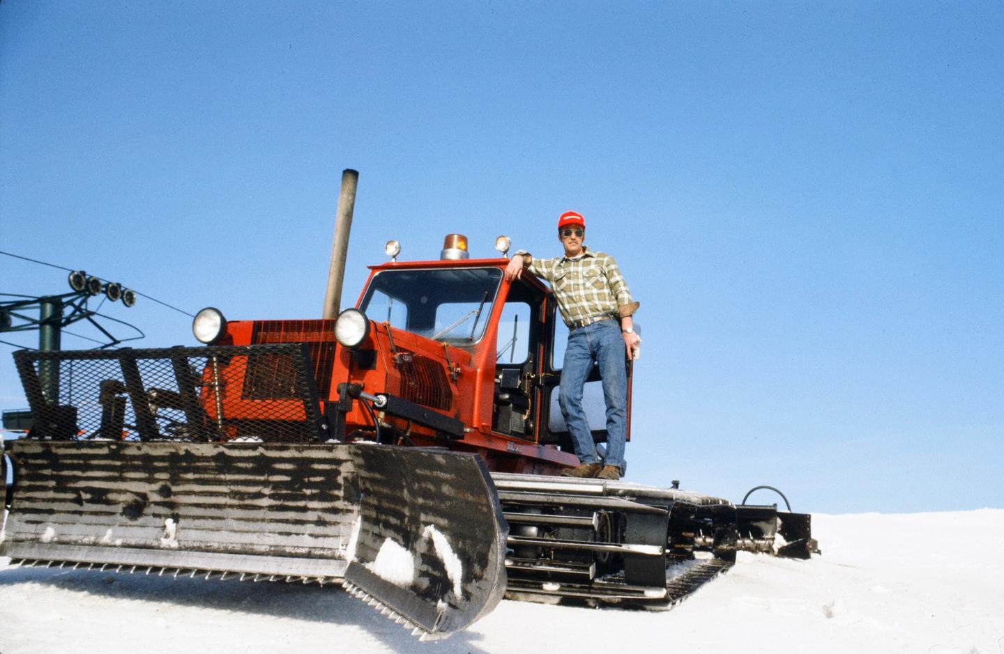Man in a red hat standing on a snow grooming machine against a blue sky.