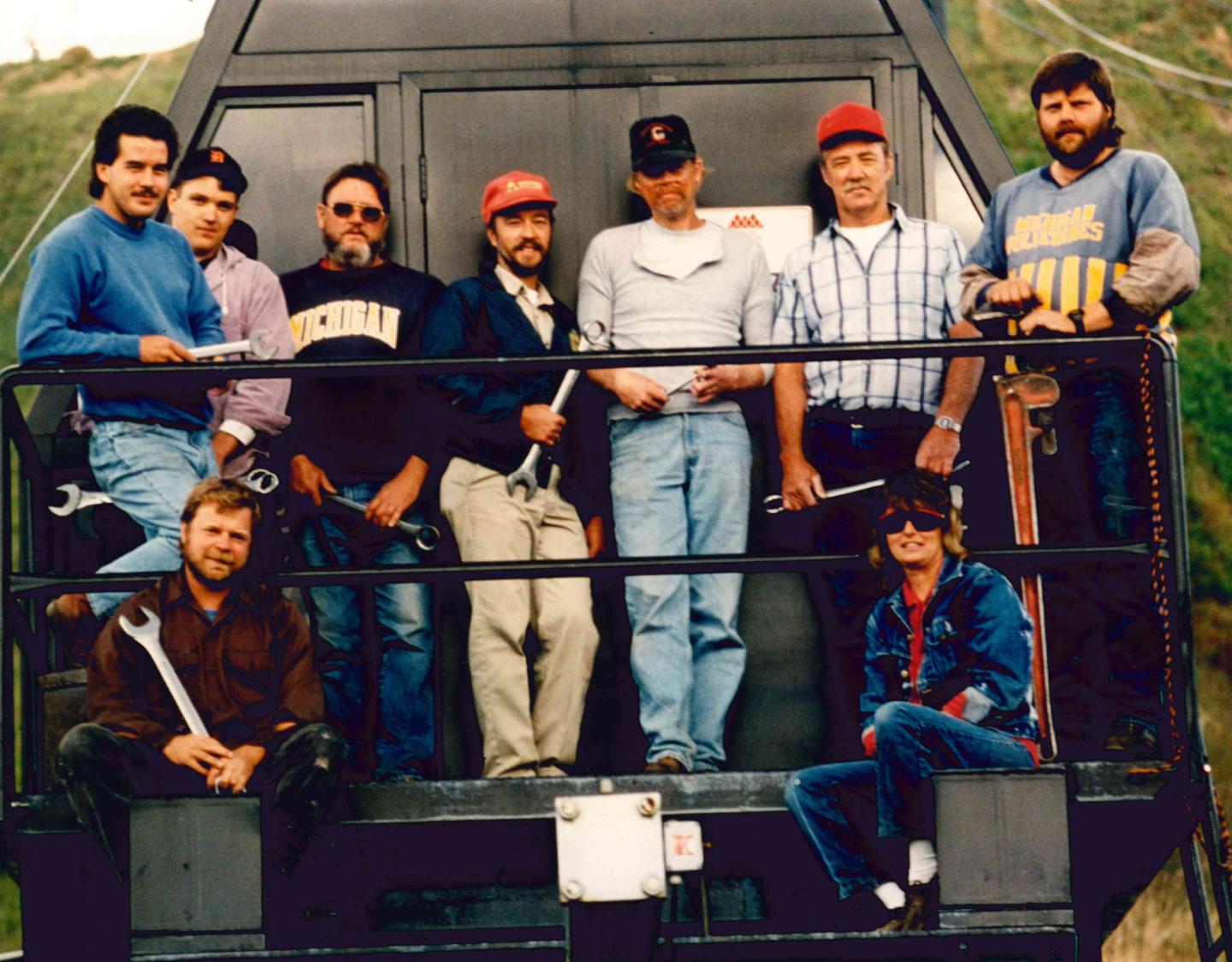 Group of men posing on an outdoor platform, some holding tools, wearing casual clothes and hats.