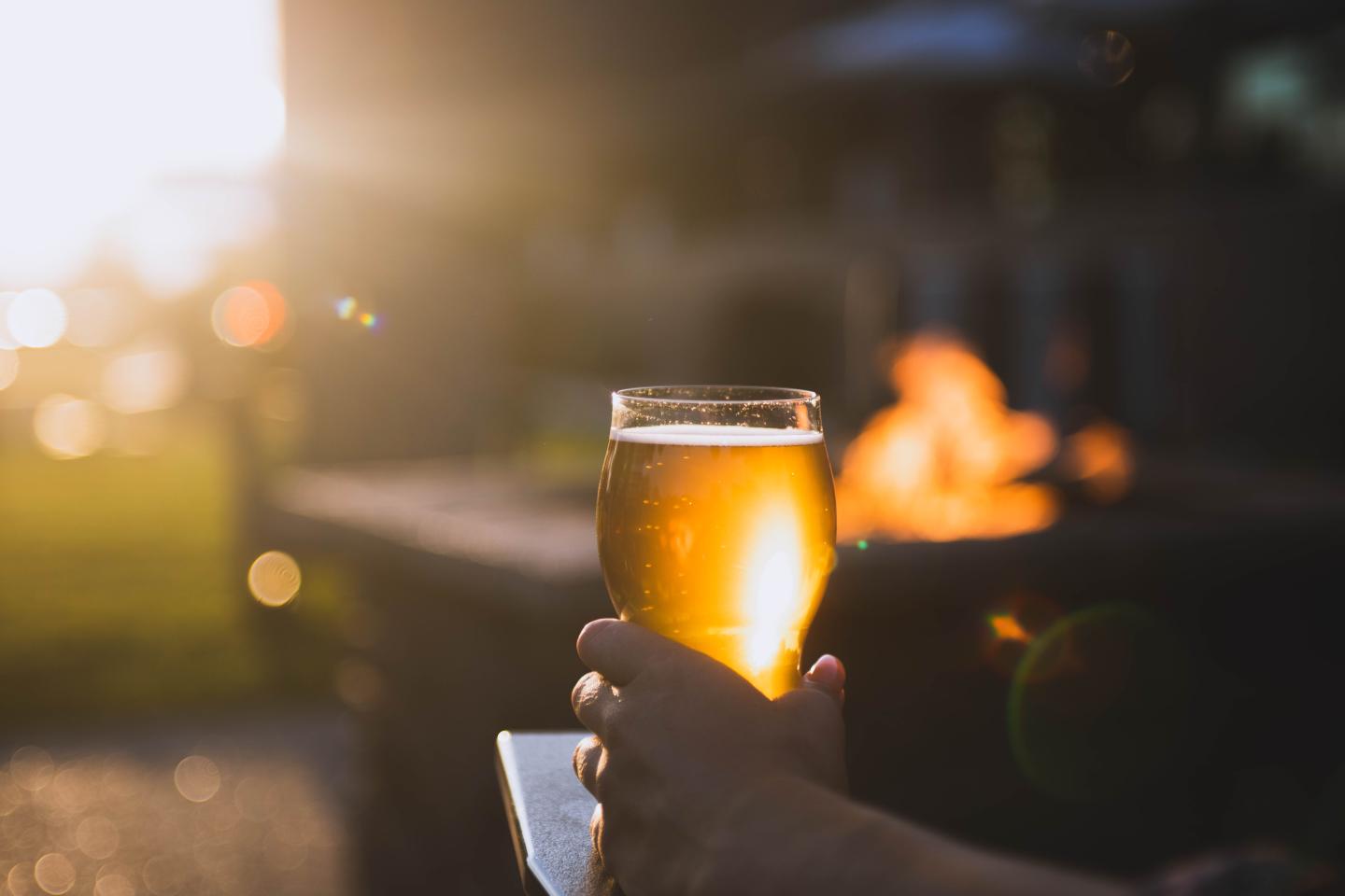 A hand holds a beer glass by a fire pit at sunset.