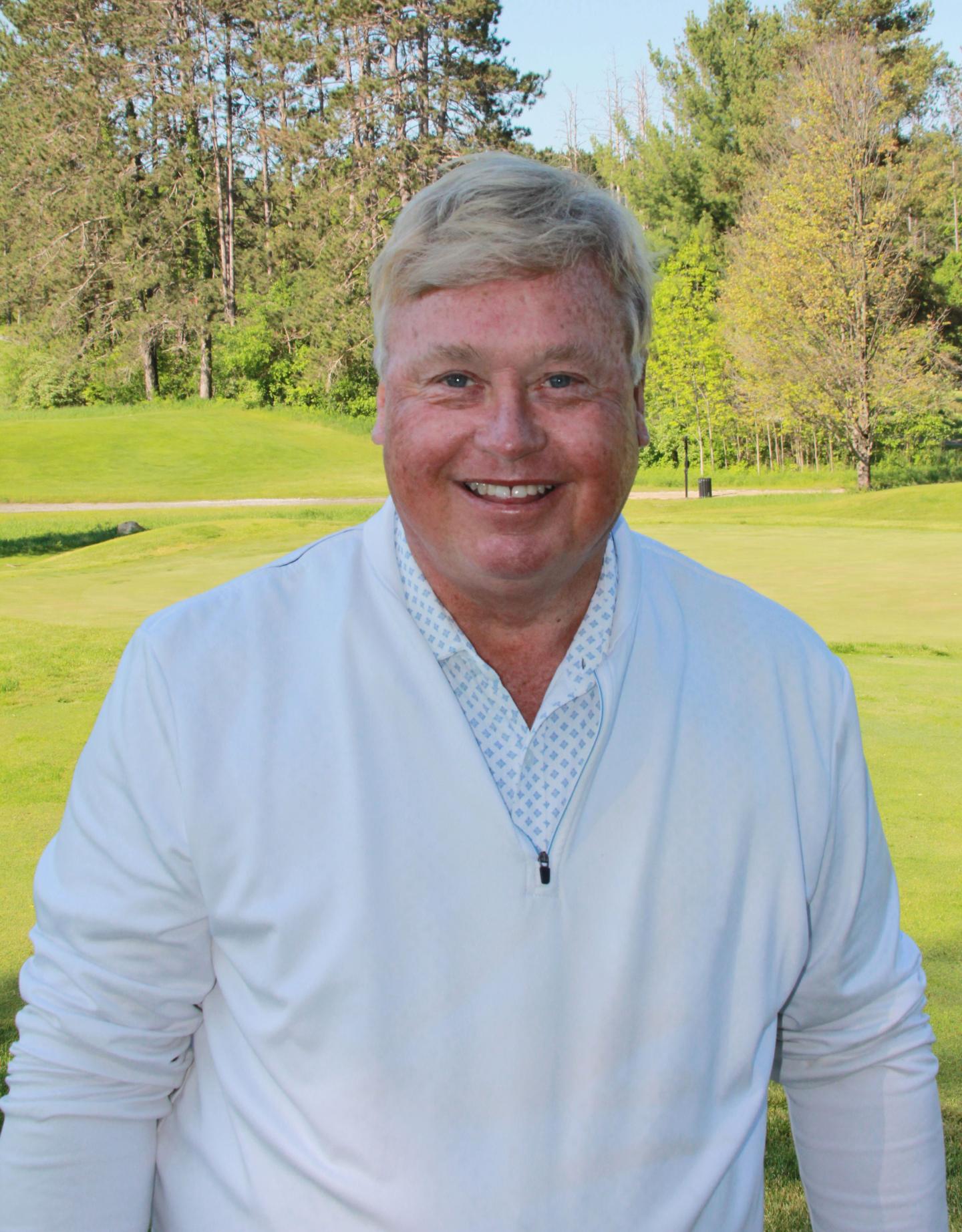 Smiling man in a white sweater stands on a sunny golf course.