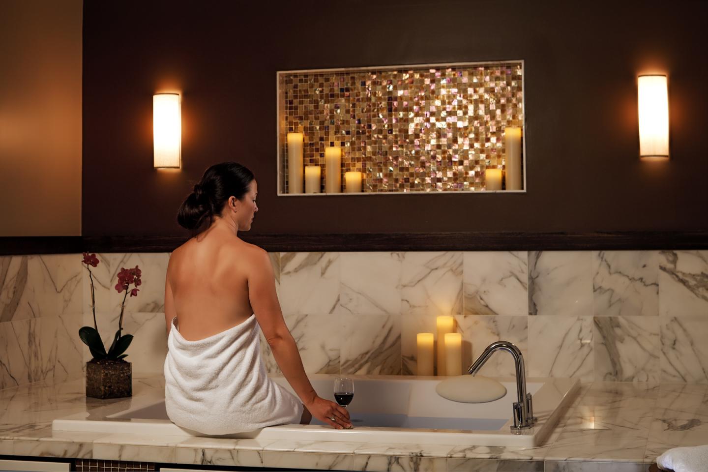 Woman in a towel sits by a marble bathtub with candles.