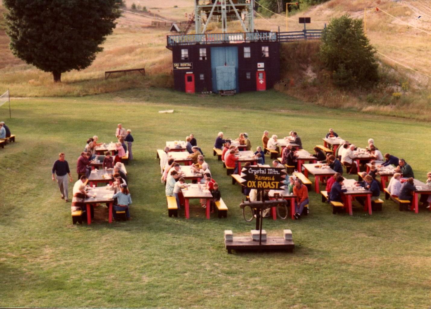 People sitting at picnic tables on grass by a building with red doors.