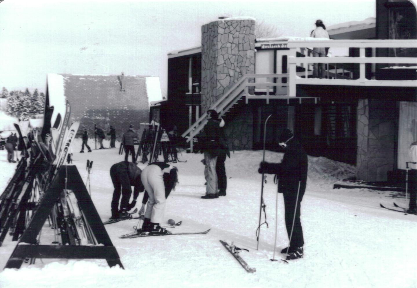 Skiers preparing on a snowy slope near a lodge, with skis and snowboards propped up.