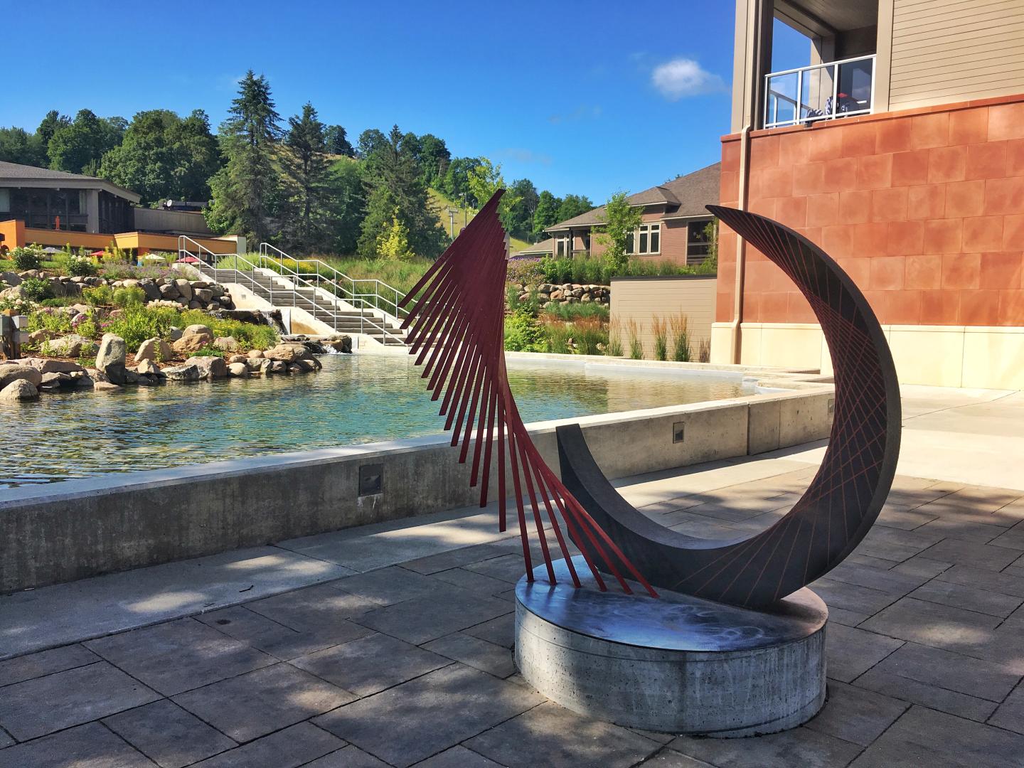 Curved metal sculpture by pond, sunny day, trees in background.