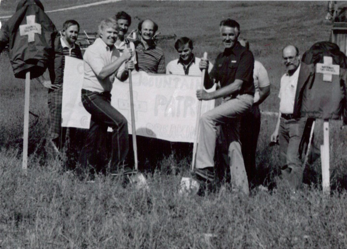 Group of men posing outdoors, holding a banner, surrounded by grassy field. Black and white photo.
