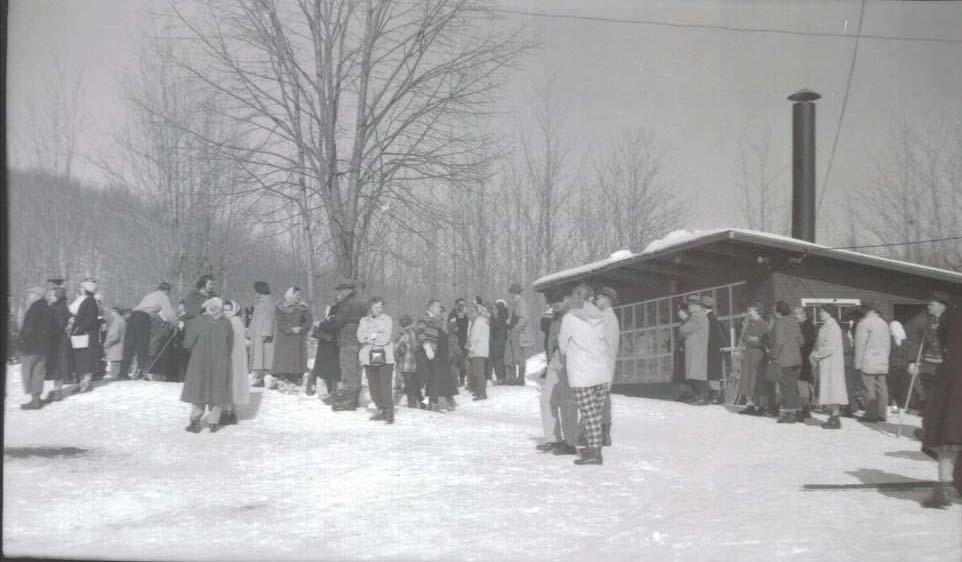 People in winter coats gathered near a snowy rustic cabin.