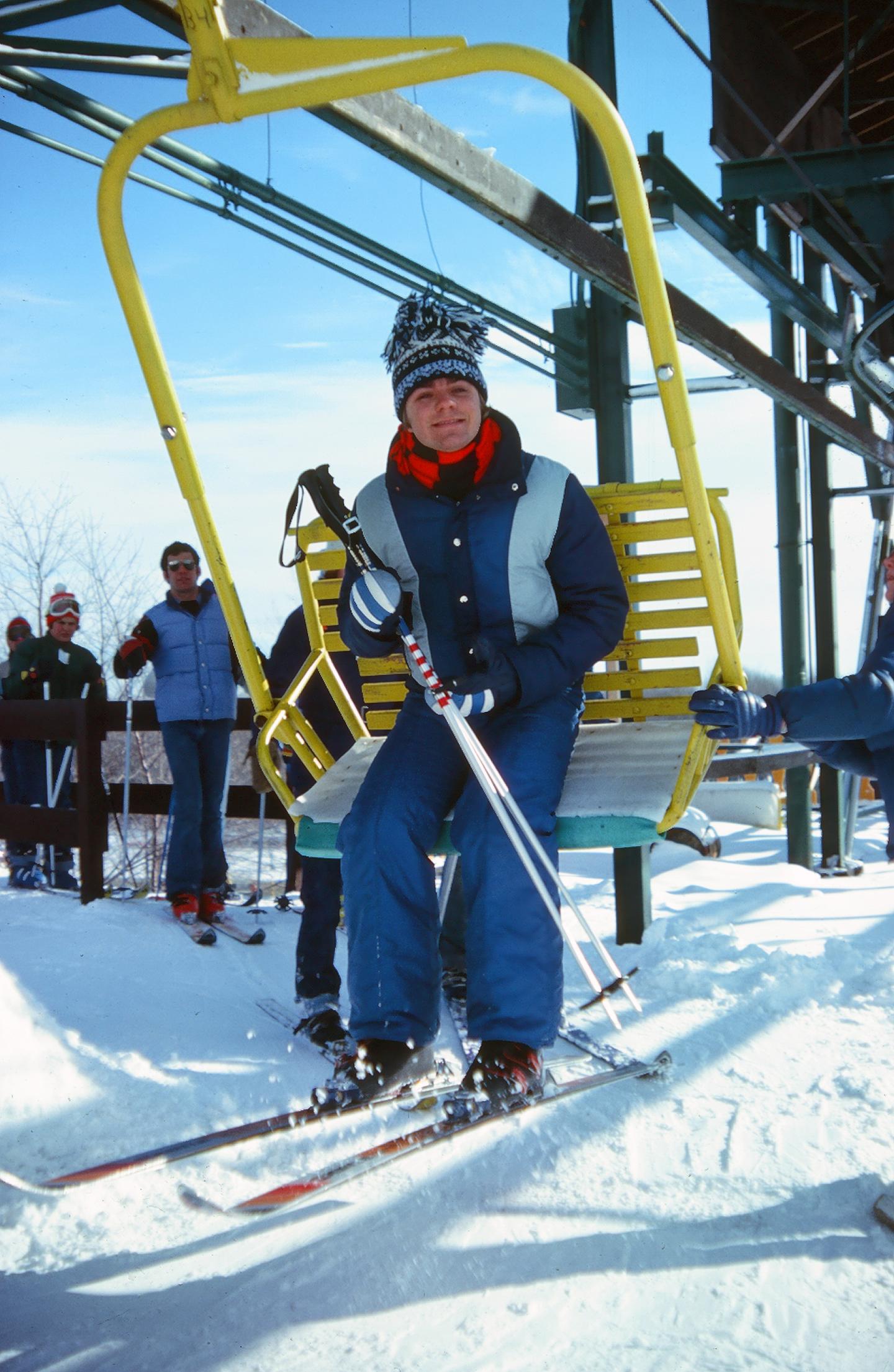 Skier in colorful gear on a yellow chairlift, clear blue sky background.