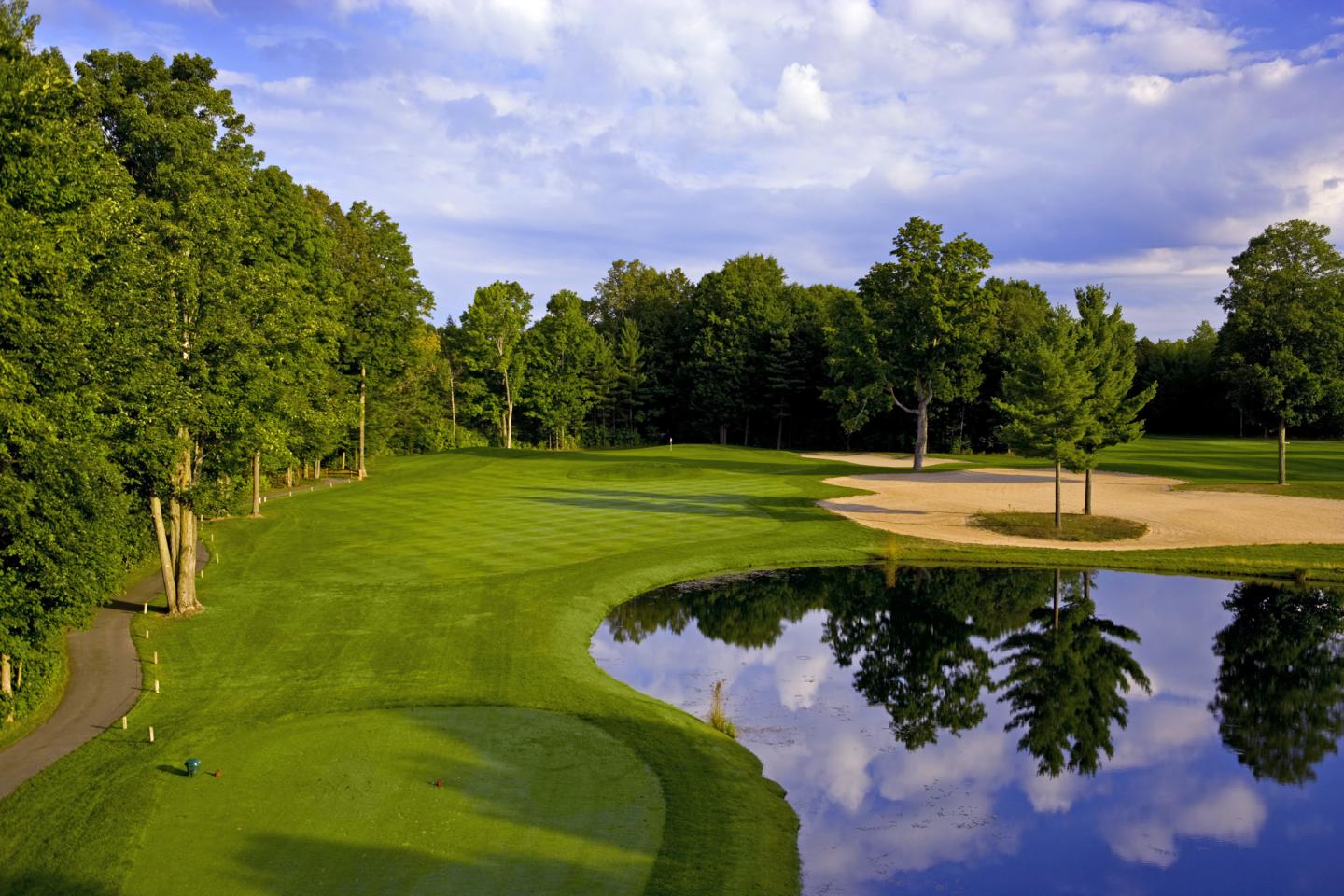 Golf course with lush greenery, trees, sand trap, and reflective pond under a blue sky.
