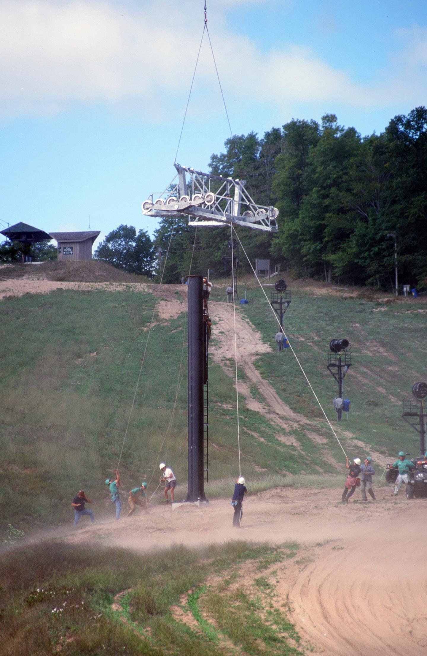 Construction workers installing a ski lift pole on a grassy hill.