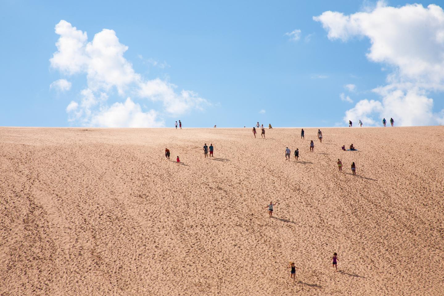 People climbing a large sand dune under a blue sky with clouds.