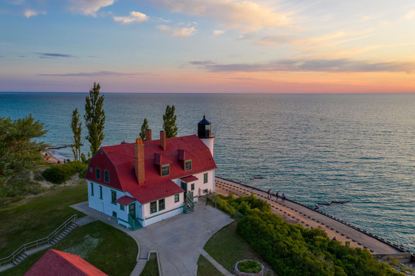 Lakeside lighthouse with red roof at sunset, surrounded by greenery.