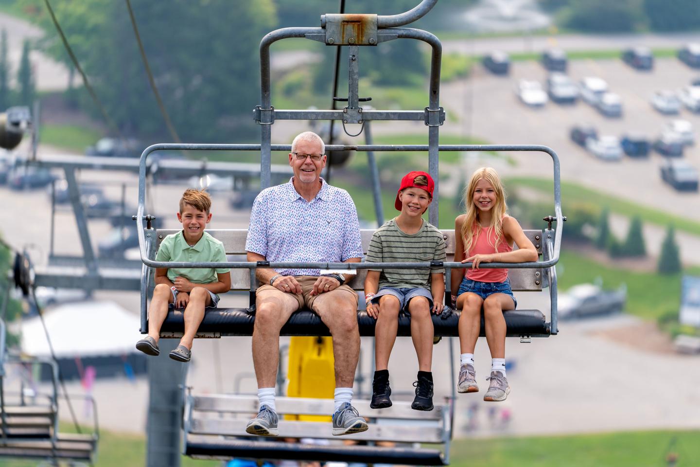 A person and three children on a ski lift, smiling on a sunny day.