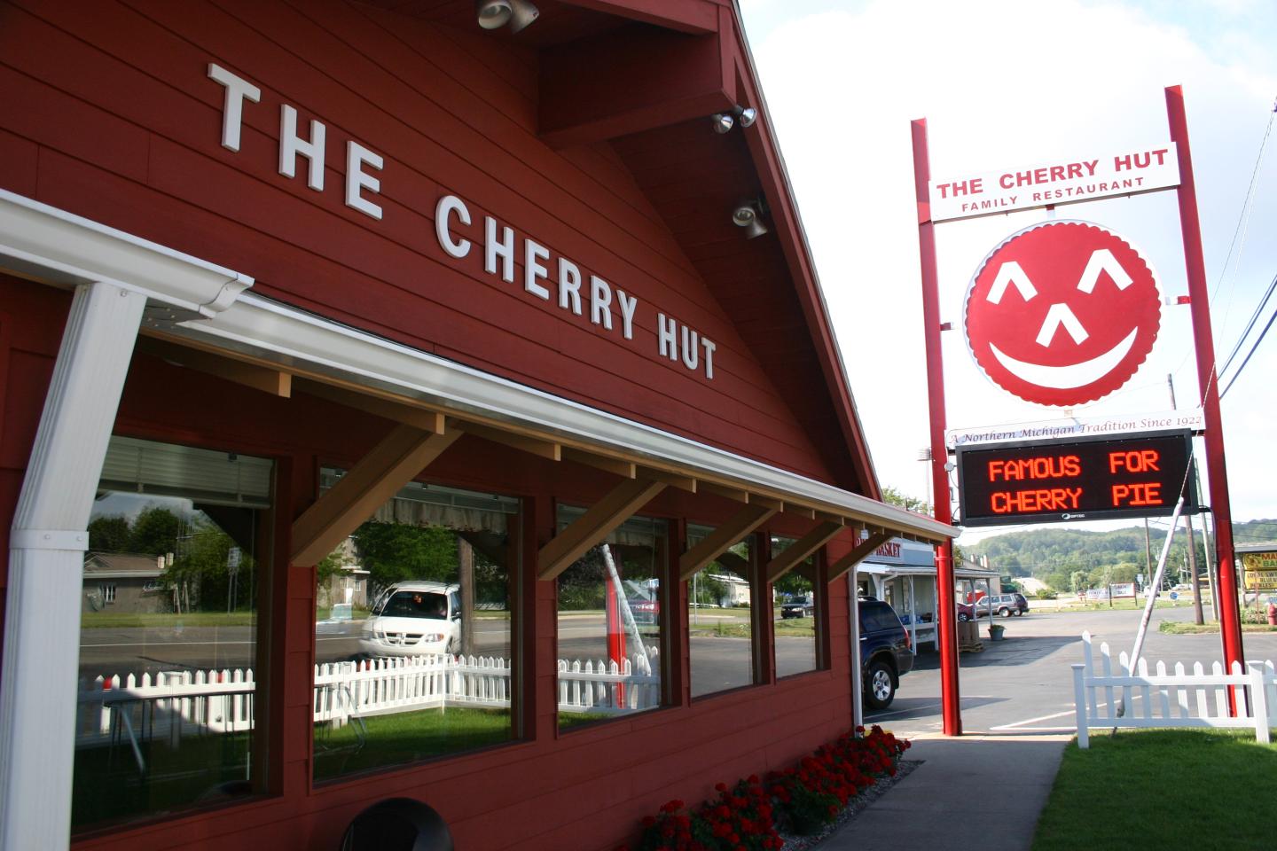 Red building with "The Cherry Hut" sign, large smiling cherry logo, and roadside in view.