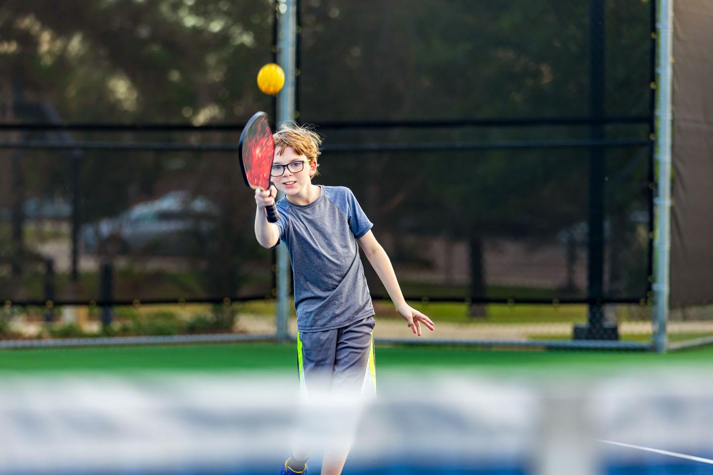 Teen playing pickleball, hitting a yellow ball with a red paddle on a sunny court.