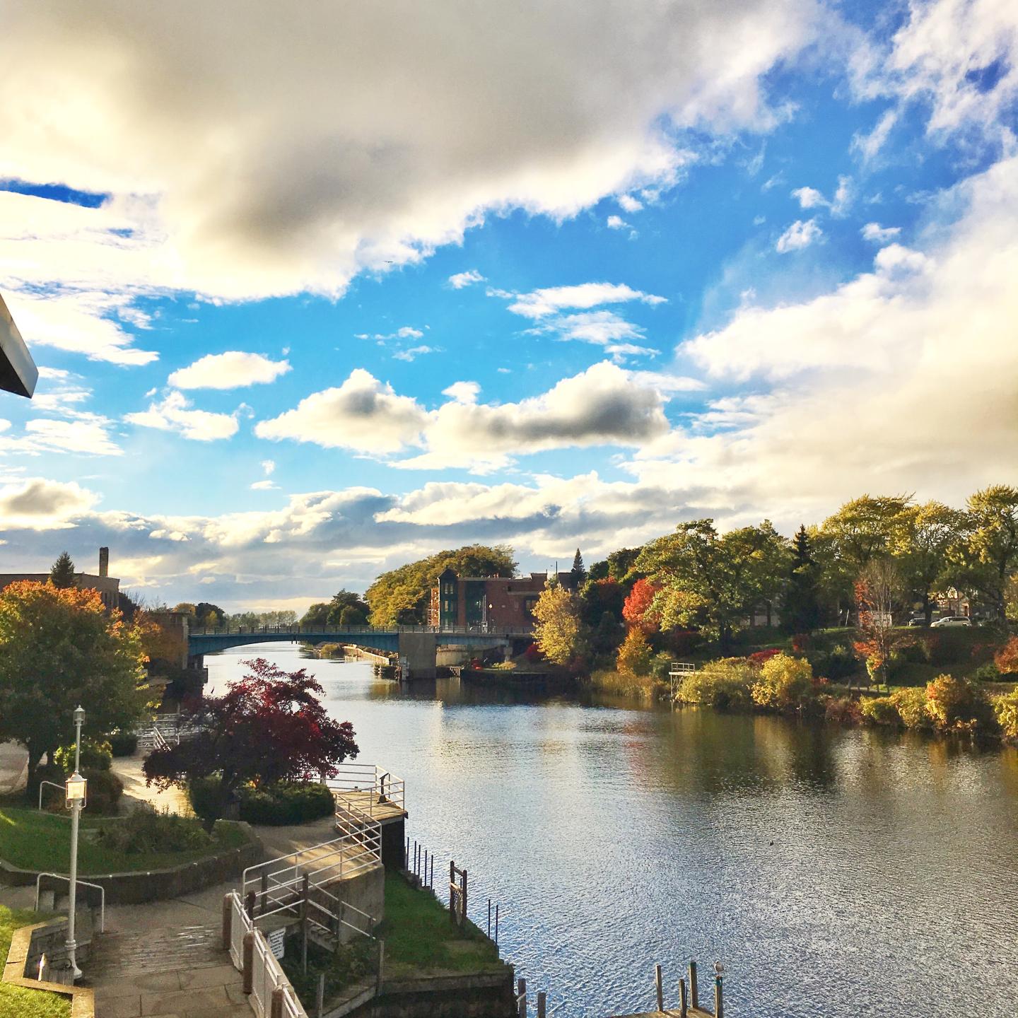 River scene with autumn trees, bridge, blue sky, and fluffy clouds.