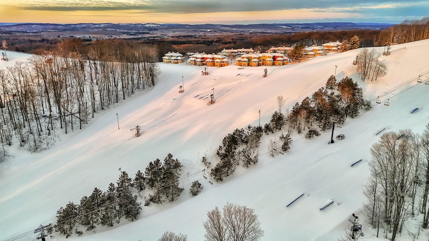Snowy ski slope at sunset with trees and distant buildings.