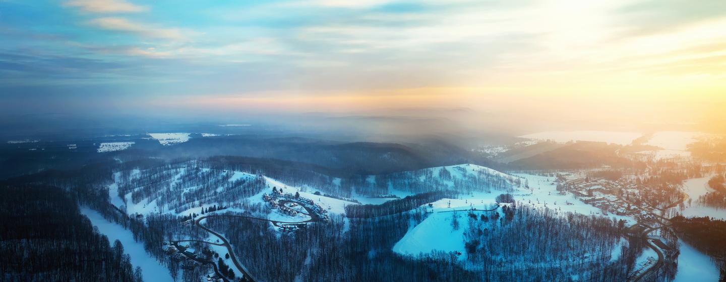 Snowy landscape at sunrise with forests and open fields, under a colorful sky.