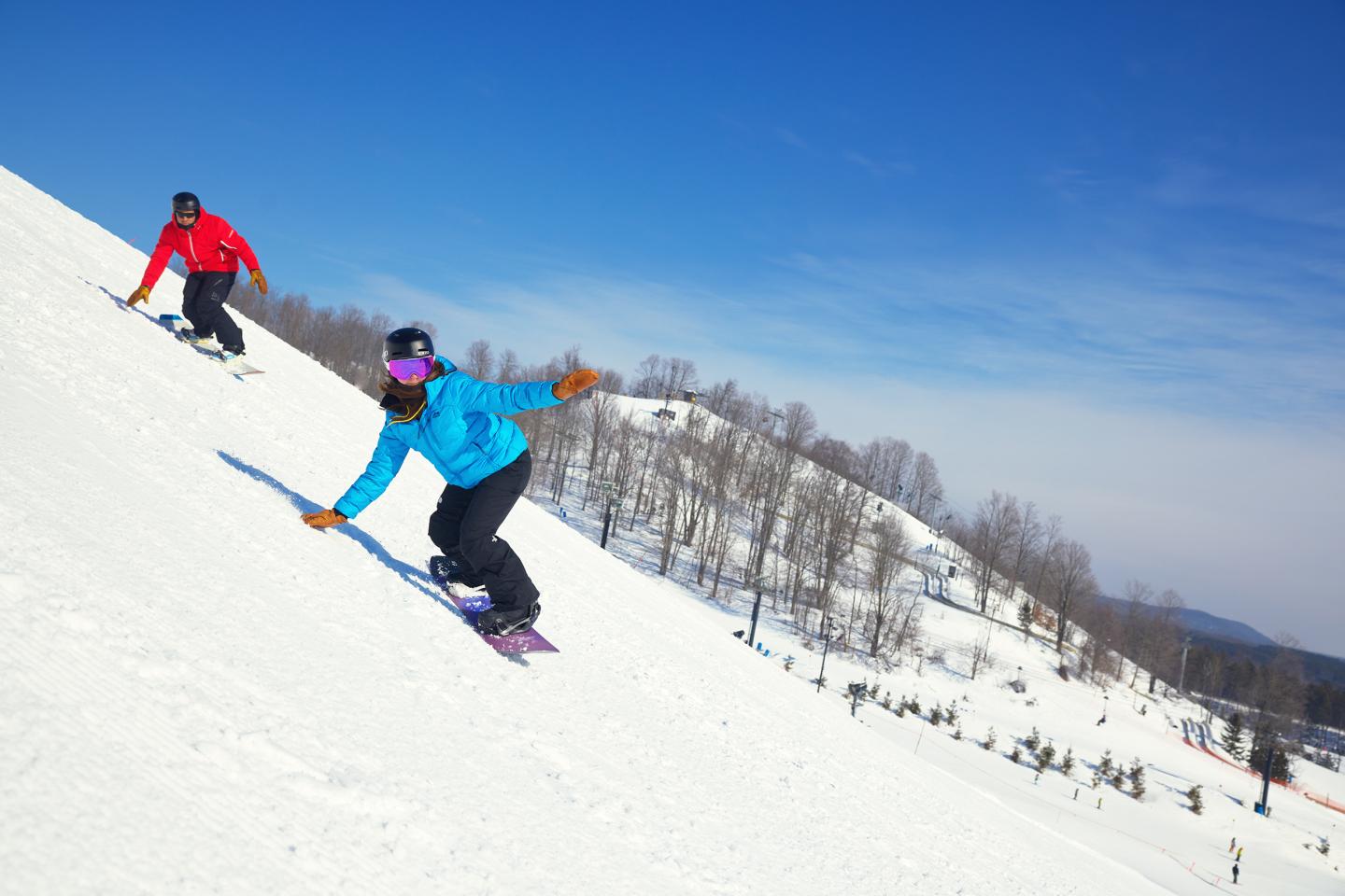 Two snowboarders descending a snowy slope under a clear blue sky.