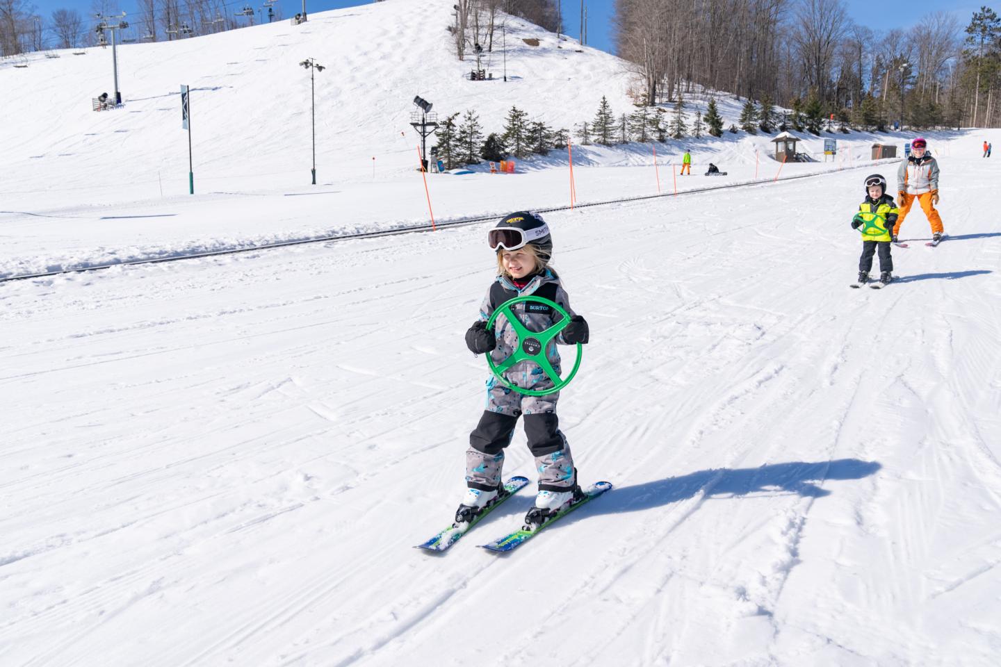 Child skiing down a snowy slope with a green harness, others in the background.