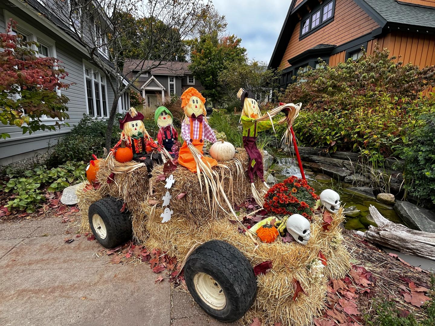 Scarecrow display with hay bales, pumpkins, and skeletons in a fall garden setting.