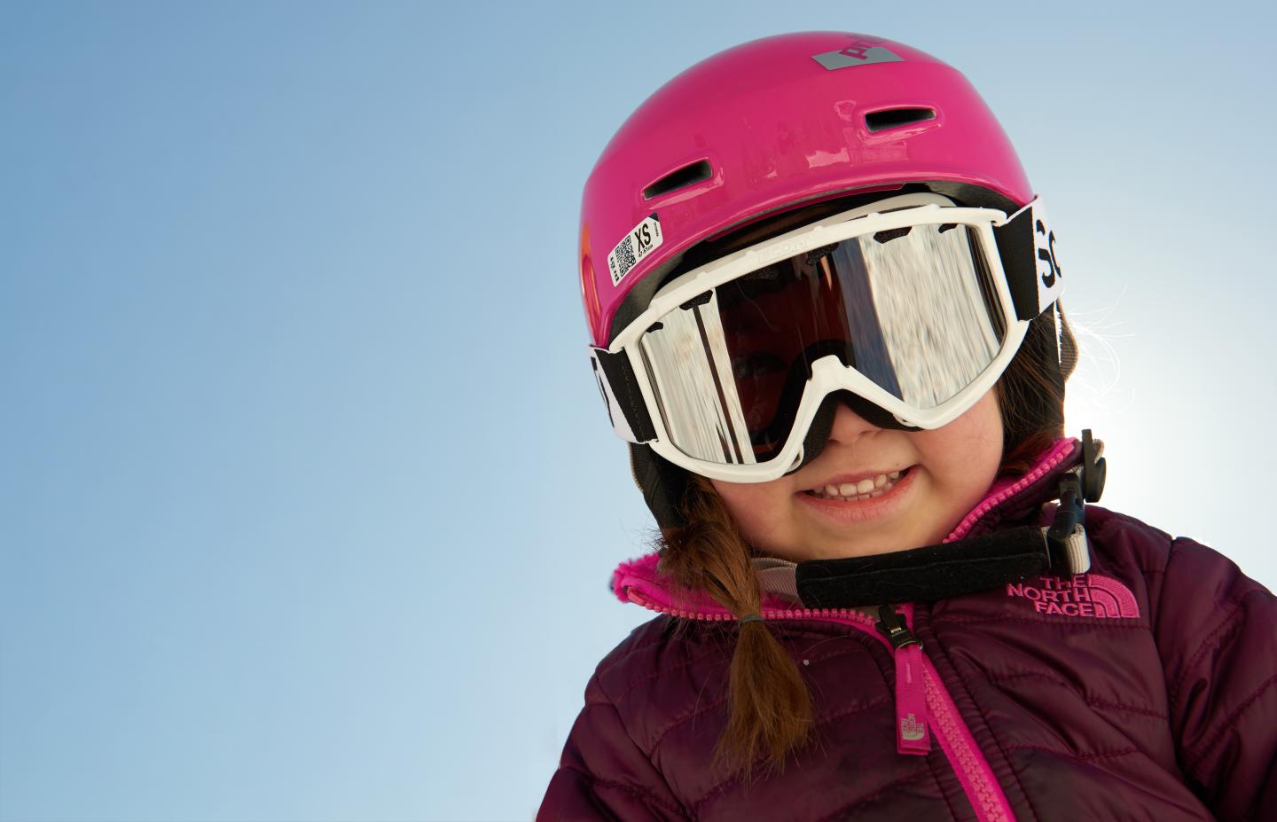 Child in a pink helmet and goggles, smiling against a clear blue sky.
