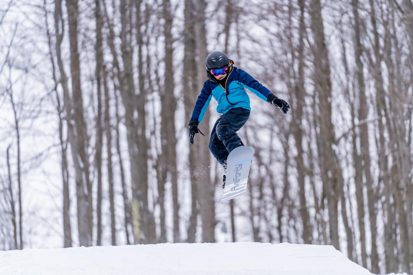 Snowboarder airborne in a forest, wearing blue and black gear.