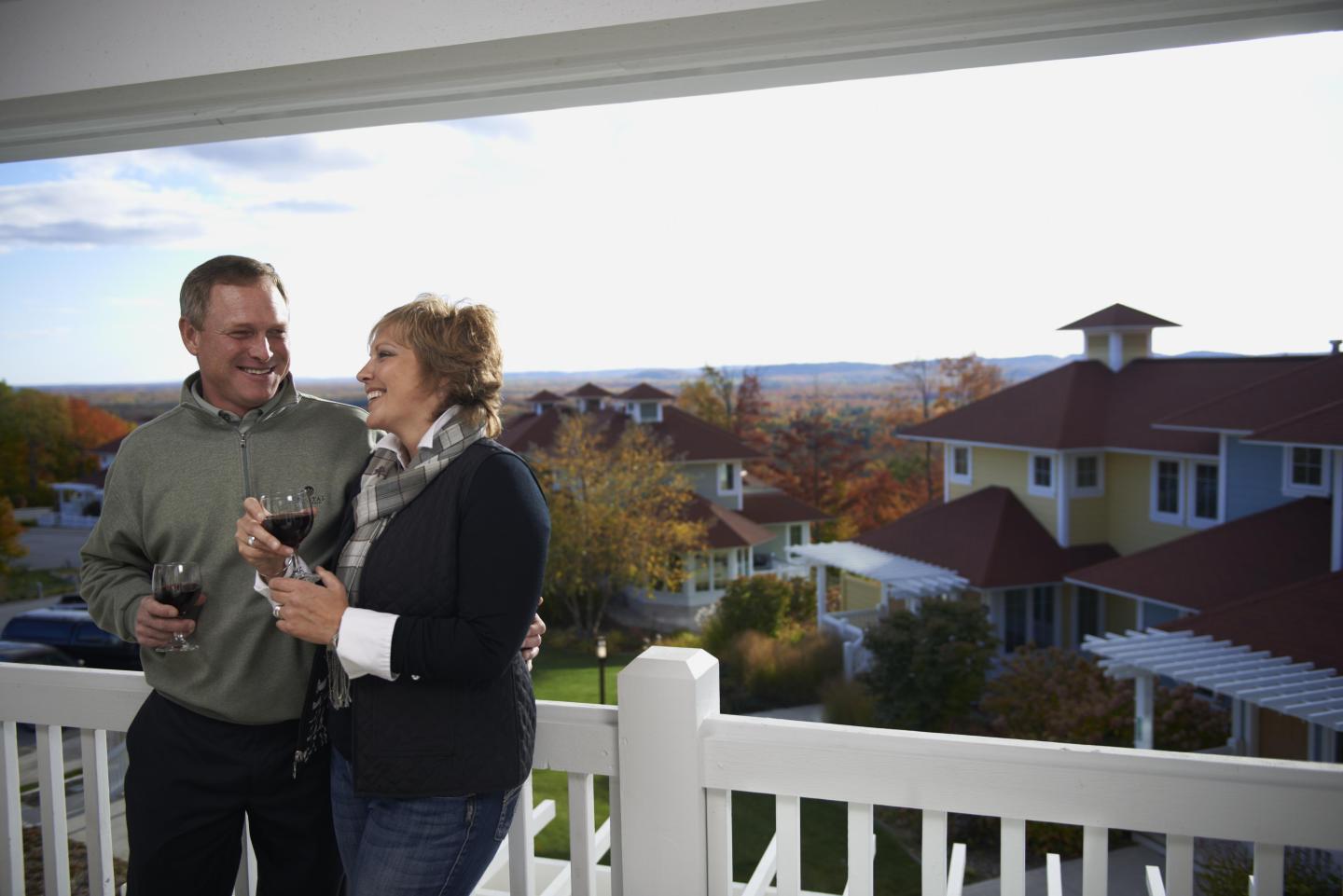 Couple holding drinks on a balcony overlooking autumn rooftops and water.