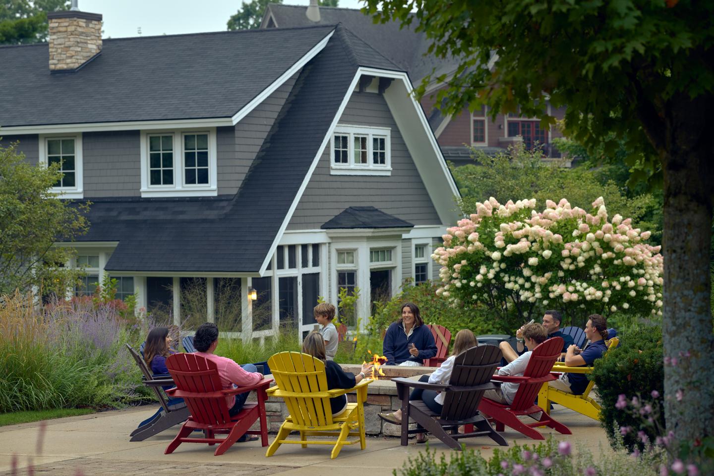 Friends relaxing in colorful Adirondack chairs outside a gray house.