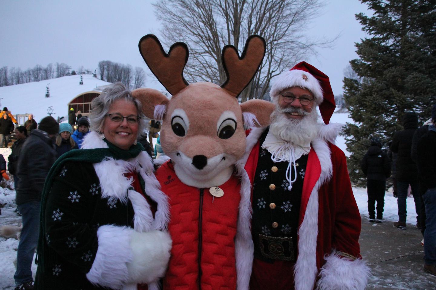 Three people in festive Christmas costumes outdoors, including a deer mascot.