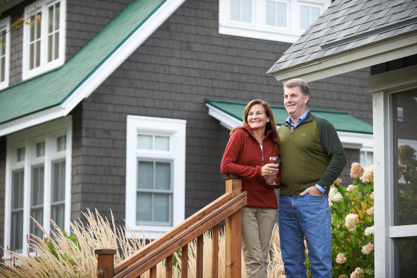 Couple standing on a porch outside a gray house, smiling together.